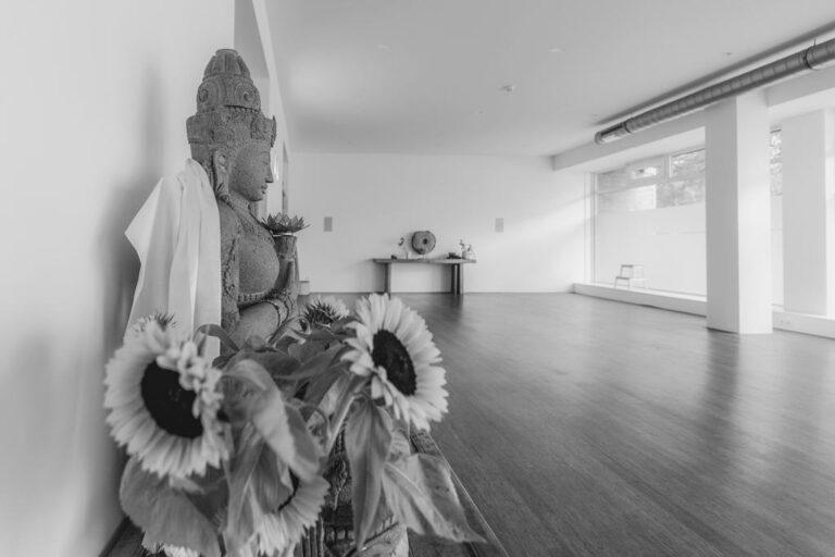 The Inner Embassy yoga studio: a calm, light-filled room with a wooden floor, a stone deity statue draped in cloth, and sunflowers by the window
