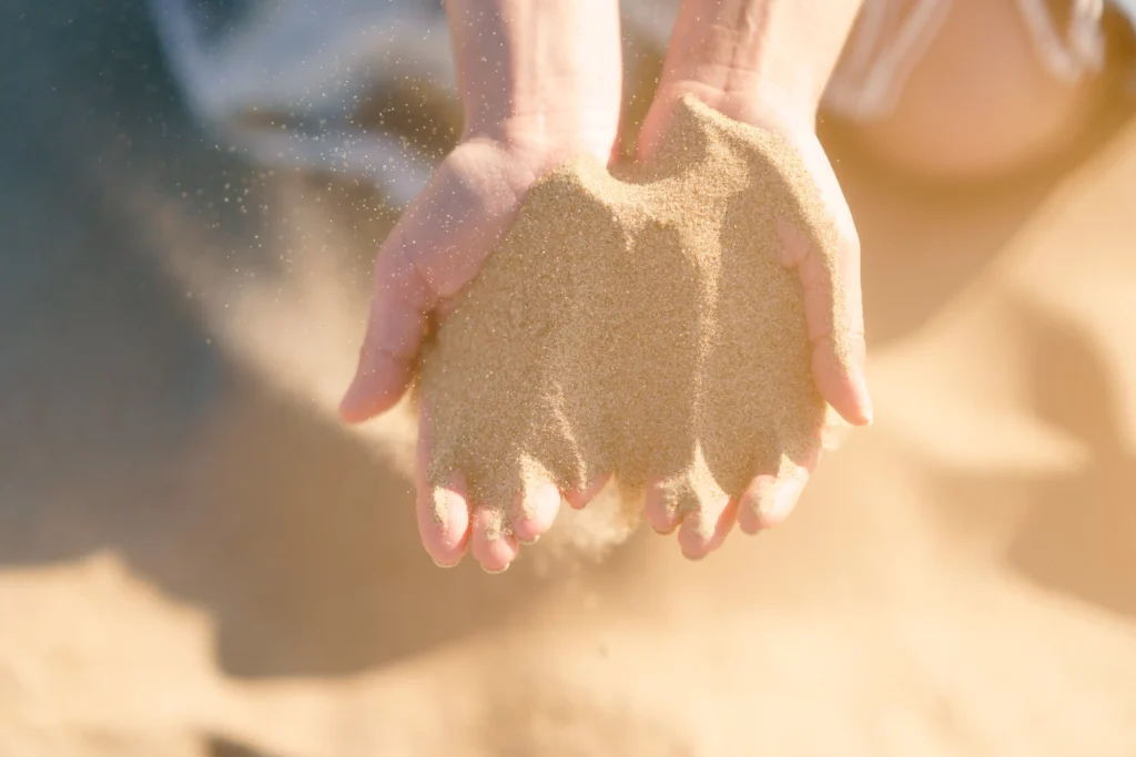 Two cupped hands letting fine, sunlit beach sand fall through the fingers