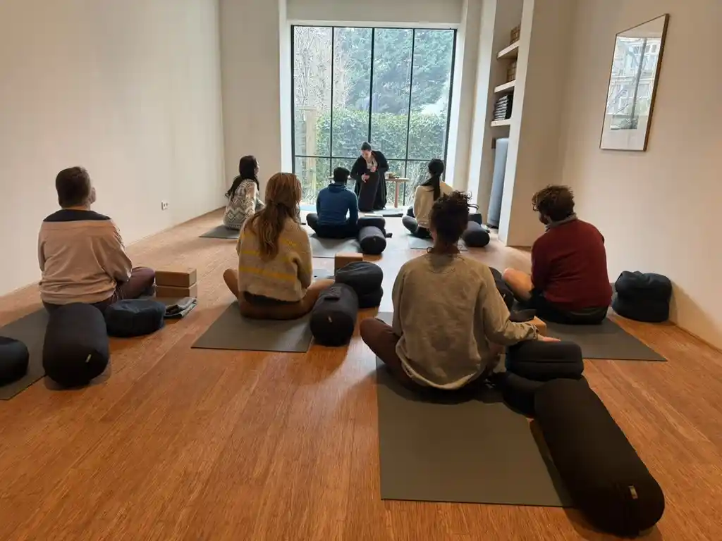 A small group sitting on mats in a circle in the light-filled Inner Embassy studio, facing Irene who leads the class from the front by the garden window